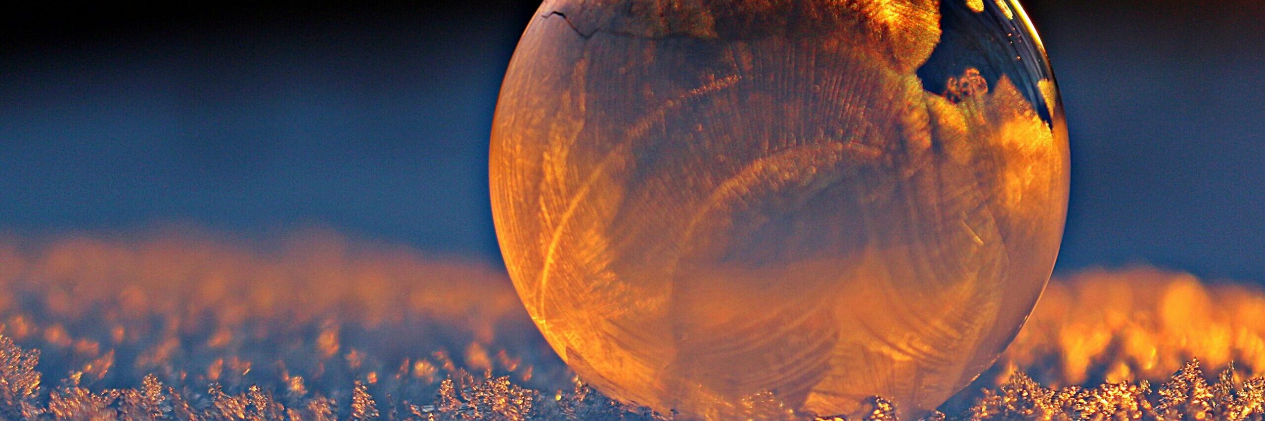 Close-up shot of a frozen bubble with warm reflections resting on a snowy surface at twilight.